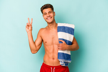 Young caucasian man holding beach towel isolated on blue background joyful and carefree showing a...