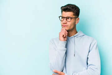 Young caucasian man isolated on blue background looking sideways with doubtful and skeptical expression. - obrazy, fototapety, plakaty