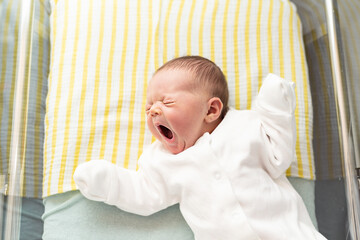 Newborn baby yawning, lying in a hospital crib, first days after birth, motherhood concept.