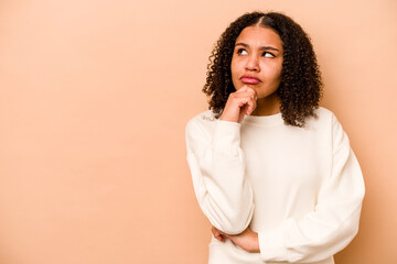 Young African American woman isolated on beige background looking sideways with doubtful and skeptical expression.