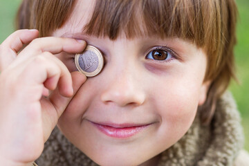 Close up portrait of toddler boy holding near eye a euro coin. Financial education concept, child and money, save and spend