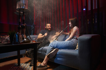 young couple smoking hookah in atmospheric hookah bar. sit freely on sofas, talking, laughing and exhaling smoke