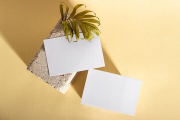 Mockup with blank horizontal paper cards with copy space on a plinth with palm leaves. Hard sunlight and shadows on a beige background. Minimal template for business layout. Top view, flat lay