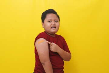 Asian boy standing and shows his arm from the vaccine. Isolated on yellow background