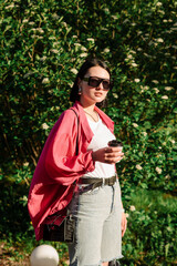 Brunette pretty woman in sunglasses holding coffee cup and walking in the park