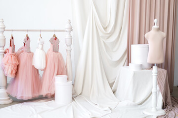 Fototapeta na wymiar Baby dresses in pink shades on a hanger next to a mannequin in a studio with fabrics. Manufacture of wearing apparel