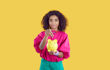 Pretty African girl holding piggybank isolated on yellow background. Child saving up money. Happy...
