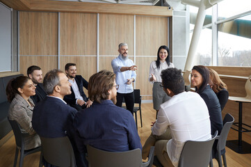 Team of cheerful business coaches talking to group of people during meeting in modern office. Successful female and male coaches speak in front of business people sitting in chairs in circle. - obrazy, fototapety, plakaty