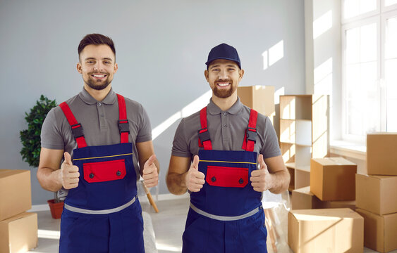 Two happy truck delivery or moving company workers guarantee best service. Portrait of young men in uniform workwear standing in living room or office, looking at camera, showing thumbs up and smiling