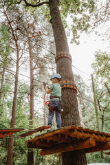 Child in sports equipment, a protective helmet stopped near a tree, holding a rope with his hands. Portrait of a brave boy climbing ropes high in tree branches. Blonde boy in Rope Park.
