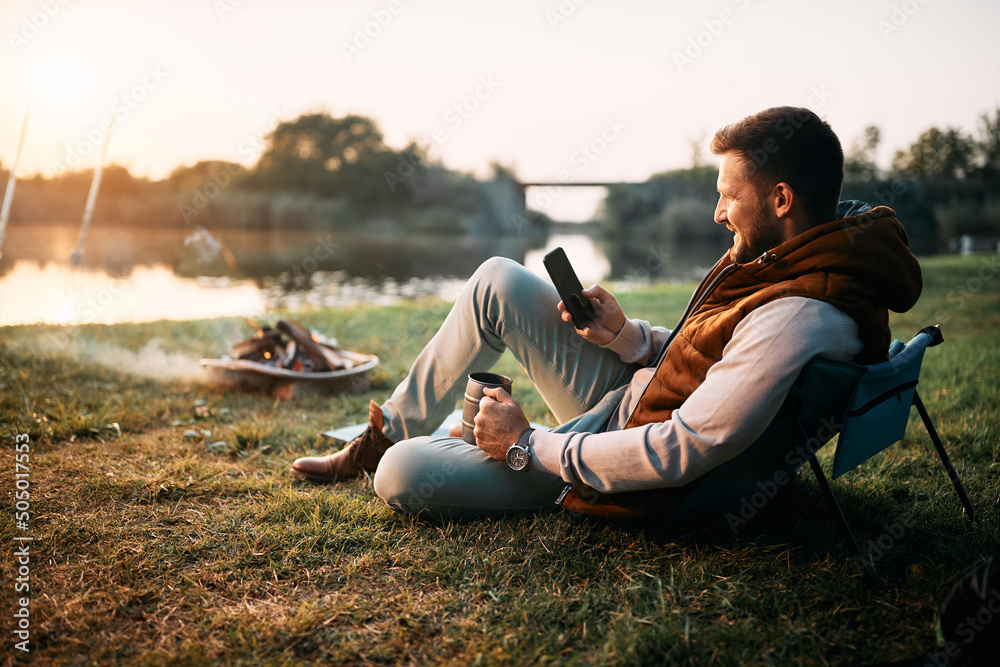 Wall mural smiling man using smart phone while relaxing by campfire in nature at sunset.