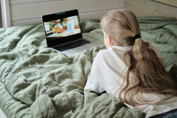 Blonde teen girl watching video on laptop while lying in bed.