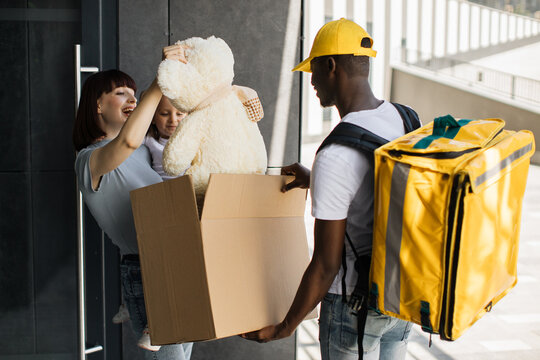 African Courier Bring Parcel To Little Girl With Mom At The Door Of Their House. Opening Parcel, Happy Child Takes Out Of Box Fluffy Teddy Bear Toy, Rejoices In The Surprise, Family Delivery.