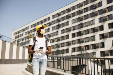 Attractive African American food delivery courier in face mask posing on the street. Smiling man in a yellow cap and with a heat-insulated food bag on his back with a phone in his hands.
