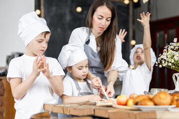 Mother and her little cute kids baking homemade sweet pie together, having fun. Home bakery, little kids in process of food preparation in the kitchen at home, helping mother, doing chores