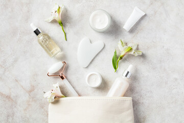 Fototapeta Makeup bag with white jade face roller, gua sha, moisturizer, serum, flowers on stone table. Skincare concept. obraz