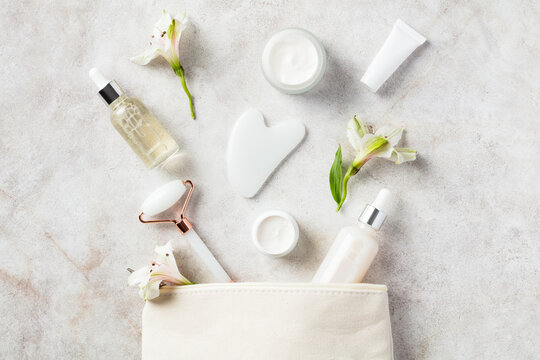 Makeup bag with white jade face roller, gua sha, moisturizer, serum, flowers on stone table. Skincare concept.