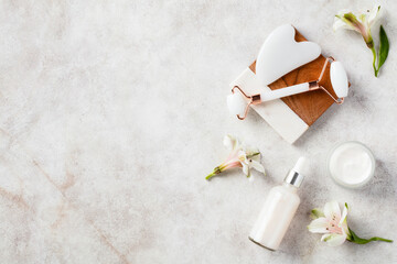 Flat lay white jade face roller, gua sha, serum, moisturizer cream, flowers on stone background. Skin and facial treatments concept.