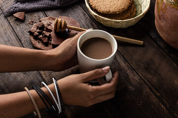 Manos de mujer sosteniendo taza de chocolate artesanal sobre mesa de madera antigua con galletas integrales