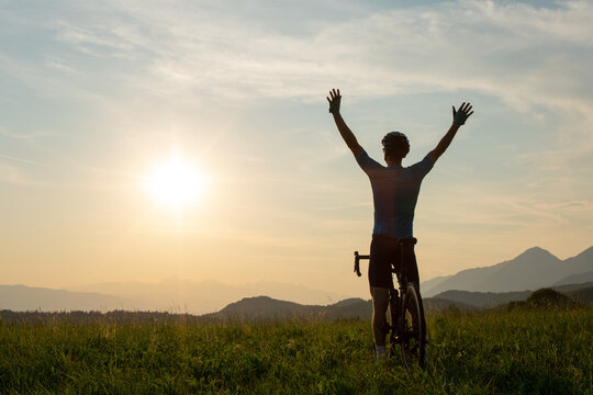 Male racing cycle rider celebrating the win with arms raised above his head, on a hill surrounded with mountains and green nature at sunset