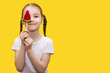 A happy girl child holds a convette on a stick in the form of a watermelon and covers part of her face with it on a yellow background. Happy child is having fun. Free space for text