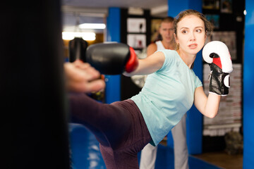 Determined athletic young woman in boxing gloves practicing heel kick by straight leg on heavy...