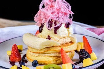 A serving of stacked pancakes with ice cream, fruit pieces and pink fairy floss