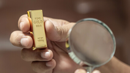 hand holding a magnifying glass and another hand holding a gold bar. gold price analysis concept.
