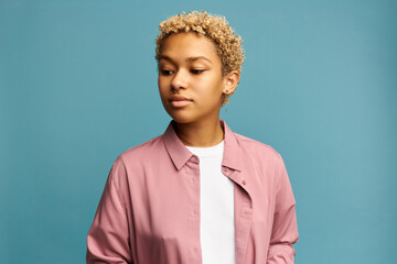 Beautiful condescending arrogant blonde African woman standing with pensive face expression, raised eyebrows, looking down, dressed in pink shirt isolated against blue background, wearing nose ring