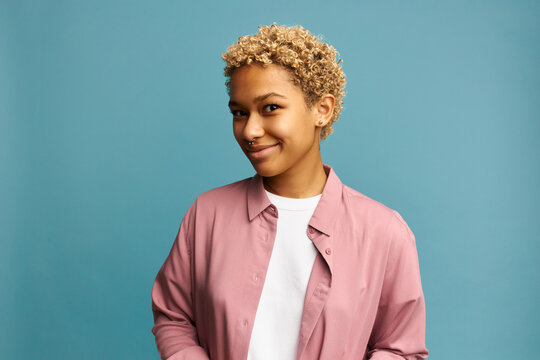 Horizontal picture of funny tricky cunning pretty blonde African American girl of 20s having sly smile, dressed in pink shirt, posing over blue wall. Human emotions and feelings. Body language