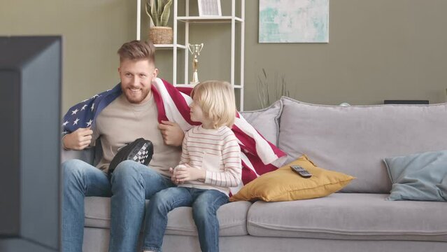 Slowmo of Caucasian man and his little son watching baseball match on television while sitting on sofa in living room with US flag and baseball equipment, celebrating winning of their team