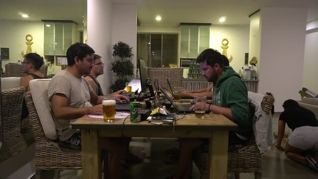 several friends playing and working with their laptops at the dining room table drinking beer and their girlfriends and girlfriends playing games on the living room floor