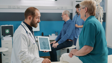 Male osteopath showing human skeleton illustration on tablet, explaining spinal cord and bones injury at exam appointment. Medical specialist having conversation with senior patient.