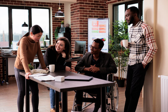African american manager with physical disability laughing with coworkers, working in disability friendly office. Businesspeople and wheelchair user chatting on break in startup workplace.