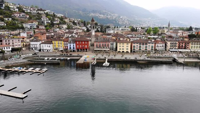 Aerial flyover along the shore of Lago Maggiore and promenade of Ascona in Ticino, Switzerland with a turn over the city rooftops revealing the lake and mountains behind the church tower