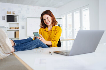 Laughing young woman sitting at her desk in office. Feet on the table and looks at her cell phone