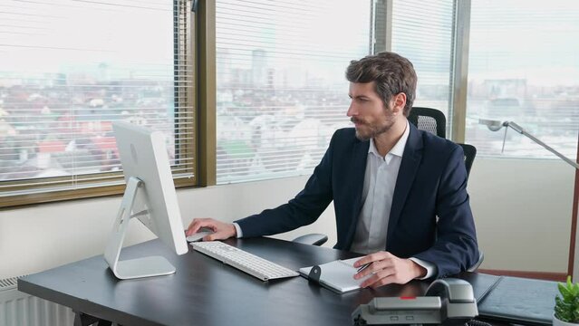 Businessman working at a computer sitting at work desk, workaholic at work, manager writes the information in a notebook, white office interior.