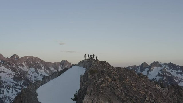Drone clip of people standing on a tall snowy apline mountain peak during sunset and surrounded by mountains