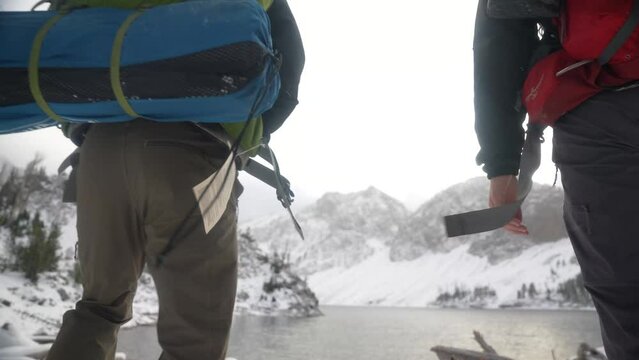 Two people backpacking and walking towards an alpine winter lake and peaks with clouds and cold temperature