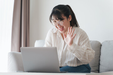 Young Asian woman using laptop computer for online video conference call waving hand making hello gesture on the couch in living room.