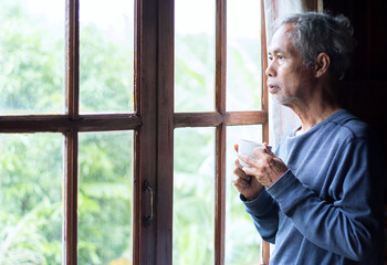 A portrait of an elderly man standing at home and looking through a window. holding a cup of coffee on a rainy day