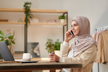 Attractive young Muslim woman wearing hijab and abaya sitting at table in her study room talking on phone