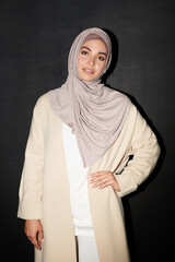 Vertical medium long portrait shot of confident Muslim girl wearing hijab standing against black...