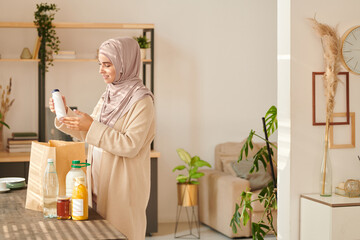 Horizontal medium long shot of cheerful young Muslim woman wearing hijab standing at table taking...