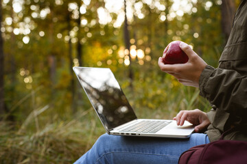Freelancer holding apple while using laptop in blurred forest 