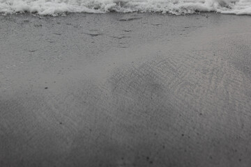 Wet black volcanic sand on seashore with splashes wave as line, texture, background.