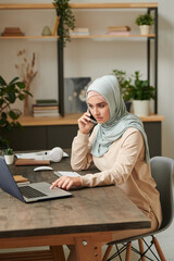 Serious young woman wearing light blue hijab sitting at table discussing important issues on phone...