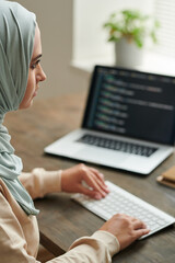 Serious Middle Eastern woman wearing hijab sitting at desk working on computer code, vertical shot