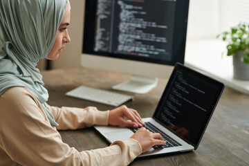 Professional programmer wearing hijab sitting at office table typing computer code on laptop, copy...