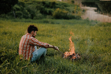 Young cheerful man staying at a campfire in the woods alone, traveler man sitting near bonfire.